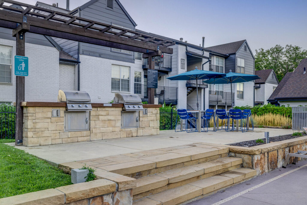 Outdoor communal grilling area with two stainless steel grills, a stone counter, bar seating with blue chairs, and blue umbrellas, located in front of a gray and white apartment building.