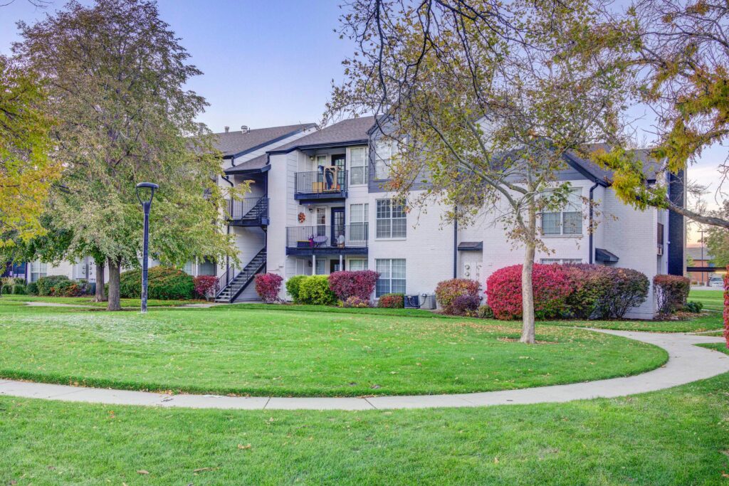 A white apartment building with black trim and exterior staircases is surrounded by green grass, trees, and colorful bushes on a landscaped lawn under a clear sky.
