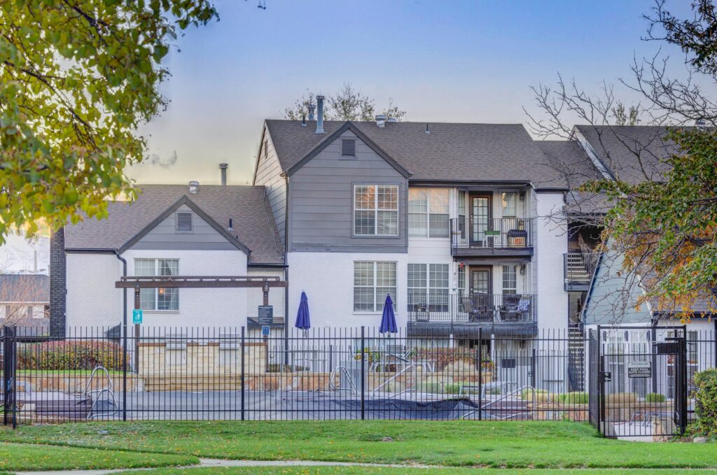 A fenced outdoor pool area with lounge chairs and closed umbrellas in front of a modern, two-story apartment building with white and gray exterior walls. Trees and grass are visible in the foreground.
