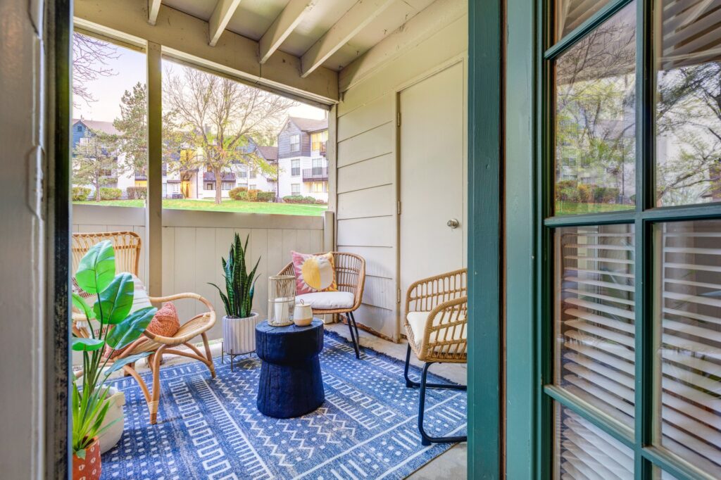 A cozy covered patio with rattan chairs, a small round black table, potted plants, and a blue patterned rug. The space is bright, with natural light and a view of neighboring houses and trees outside.