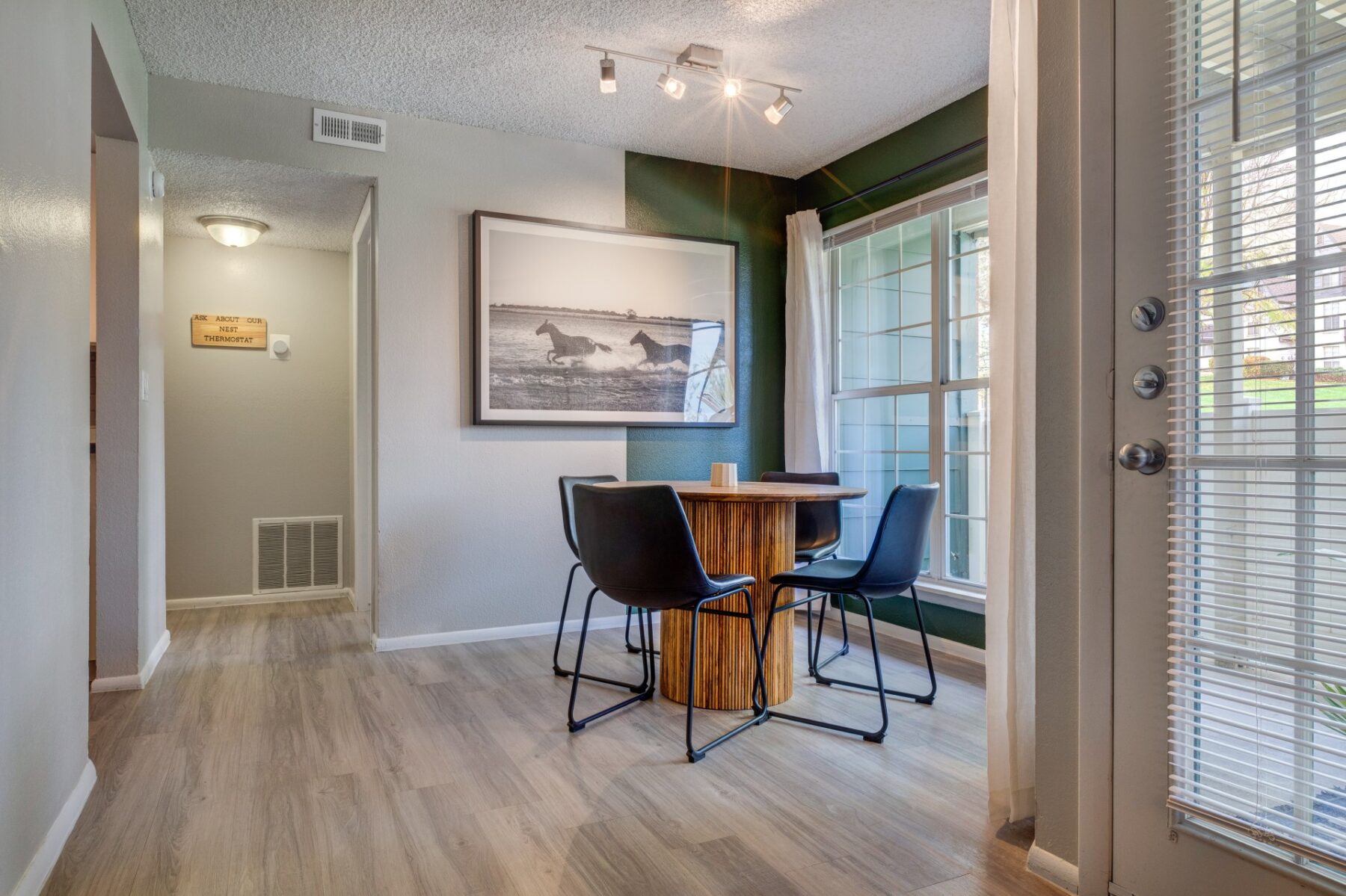 A modern dining area with a round wooden table and four black chairs, large window with white curtains, and a black-and-white horse photo on the wall. Light wood flooring and track lighting complete the space.