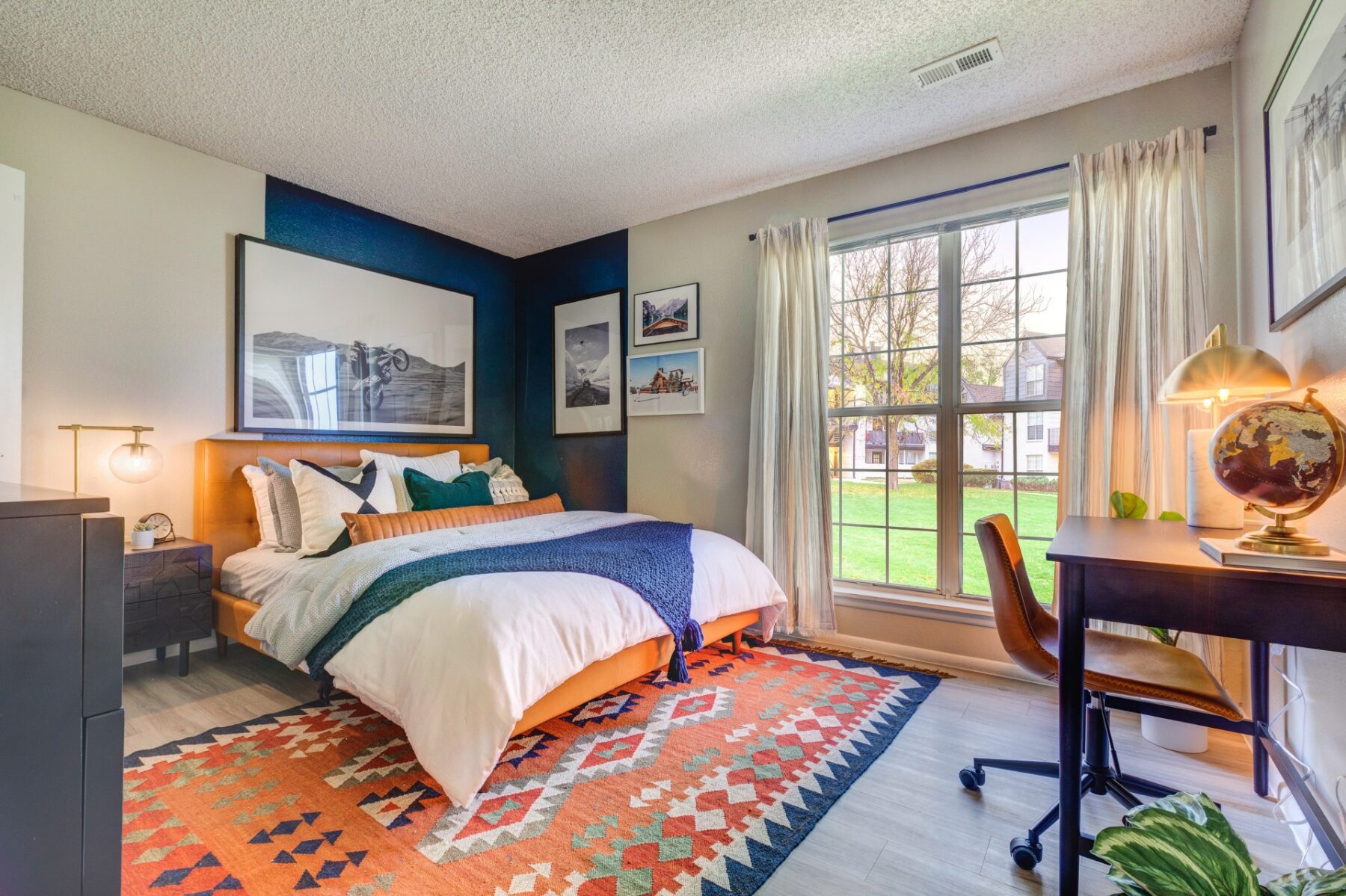 A bright, modern bedroom with a large bed, colorful geometric rug, desk with a globe lamp, and a window with white curtains overlooking a green yard. Framed artwork decorates the walls.