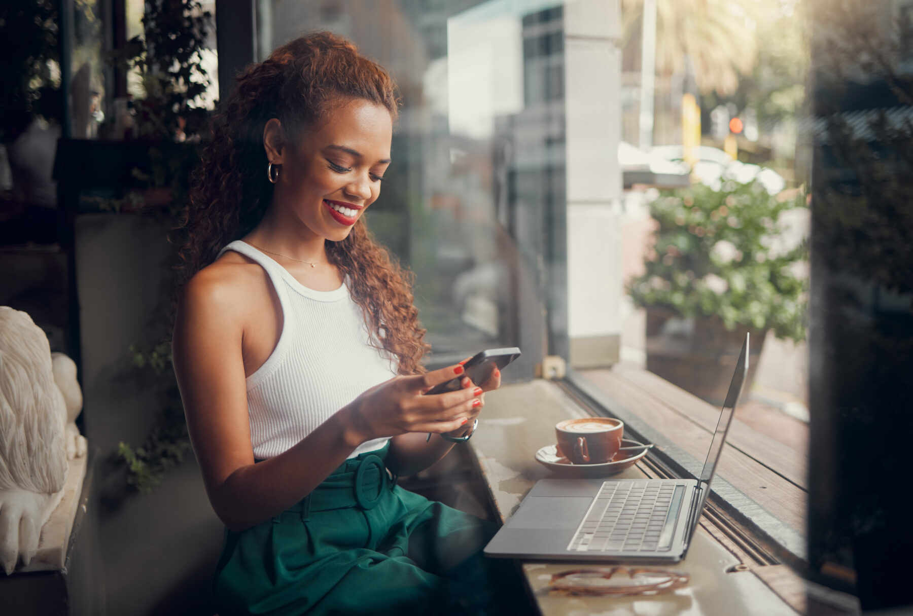 A woman sits by a window at a cafe, smiling at her phone. She has a laptop and a cup of coffee on the table, with sunlight streaming in and plants visible outside.
